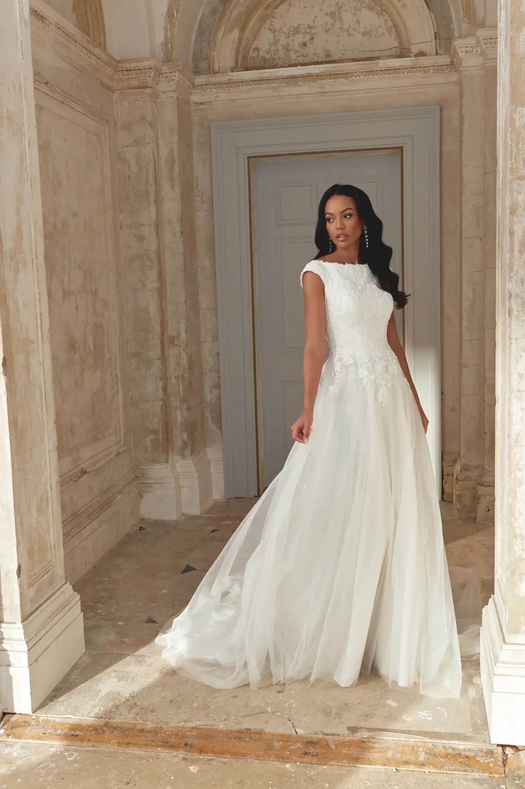 A woman wearing a flowing white dress stands in an elegant, partially ornate room with light-colored walls and a large doorway.
