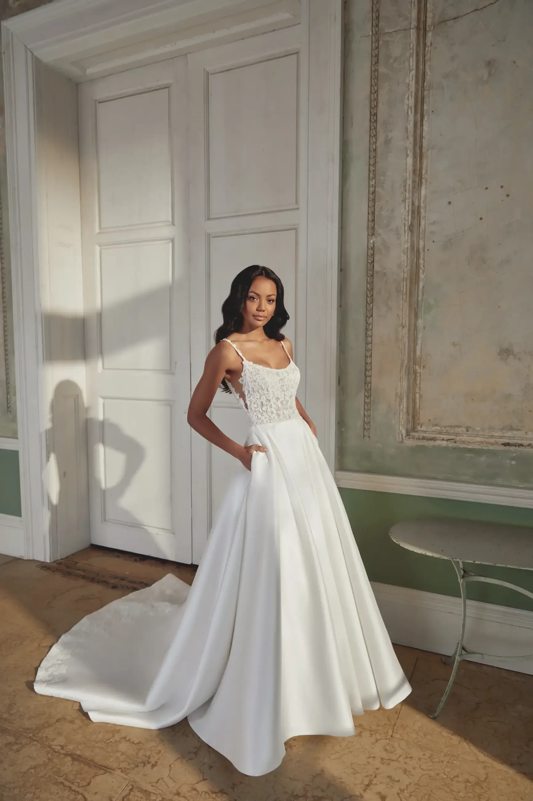 A woman stands in an elegant white wedding gown with a lace bodice and full skirt. She poses in a sunlit room with vintage decor, conveying a serene and graceful ambiance.