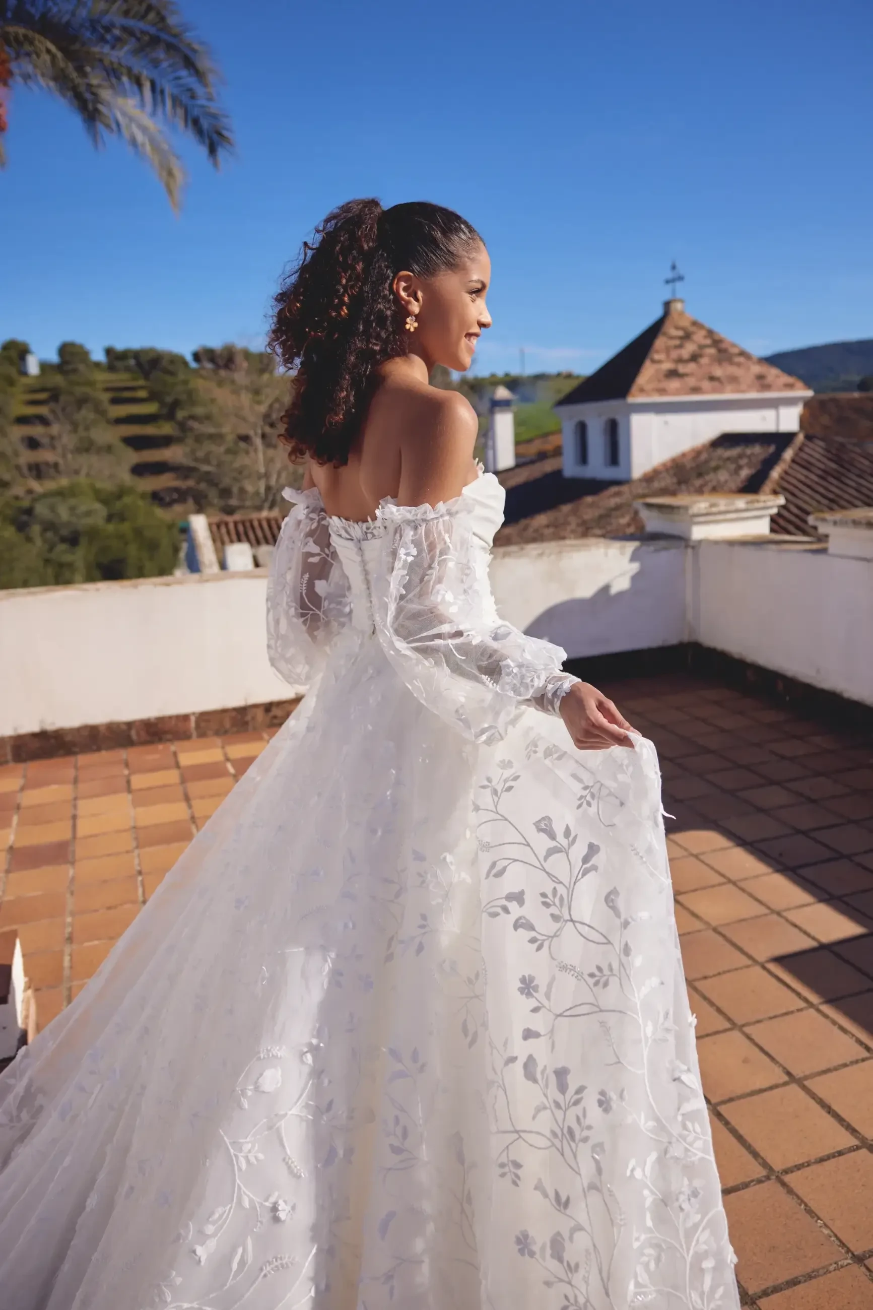 A woman in a white wedding dress with long sleeves stands on a terrace, looking back over her shoulder. The dress features floral embroidery, and the setting includes green hills and blue sky.