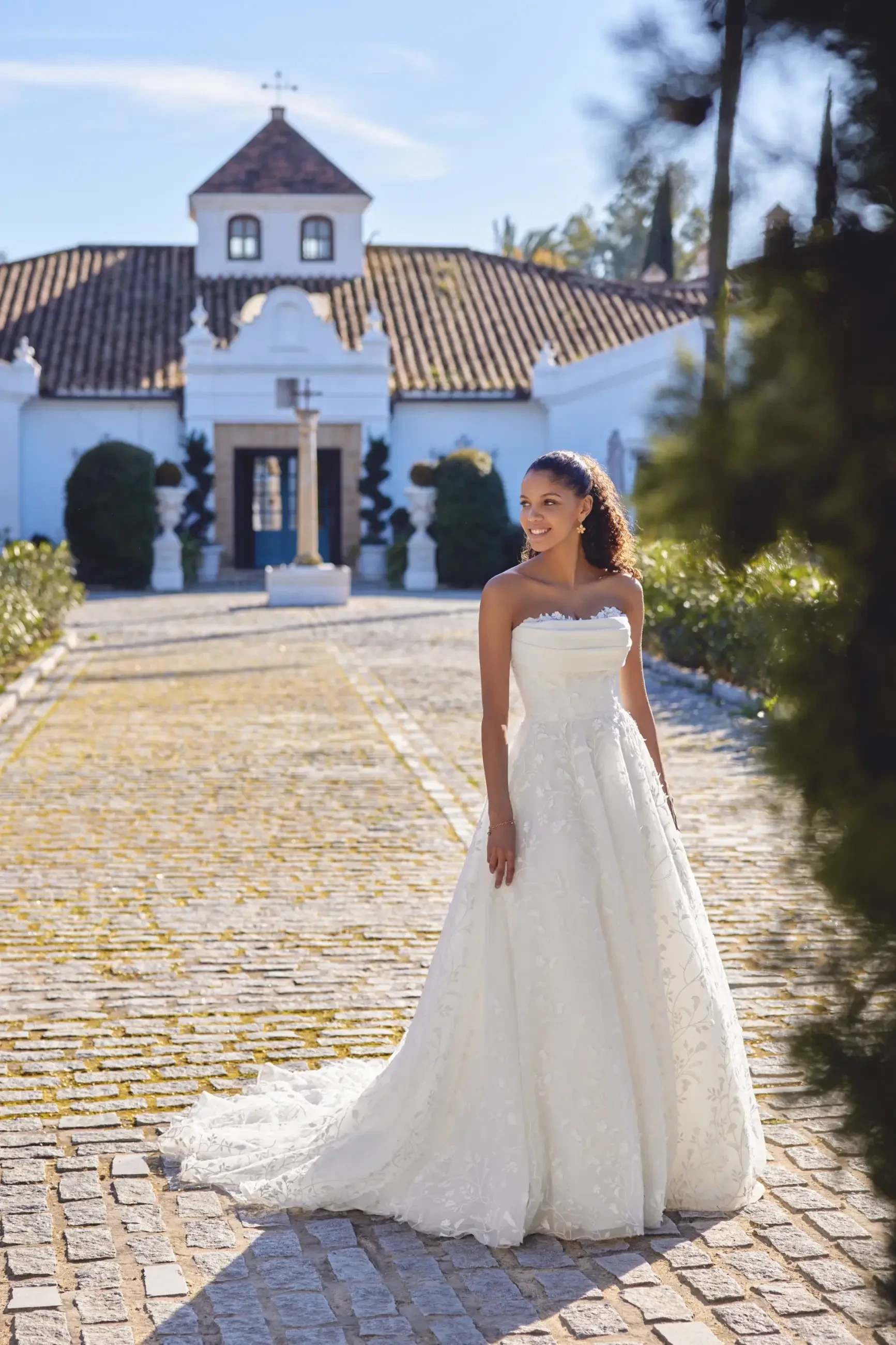 Bride in a strapless white gown stands smiling on a sunlit cobblestone path, with a picturesque chapel in the background, surrounded by greenery.