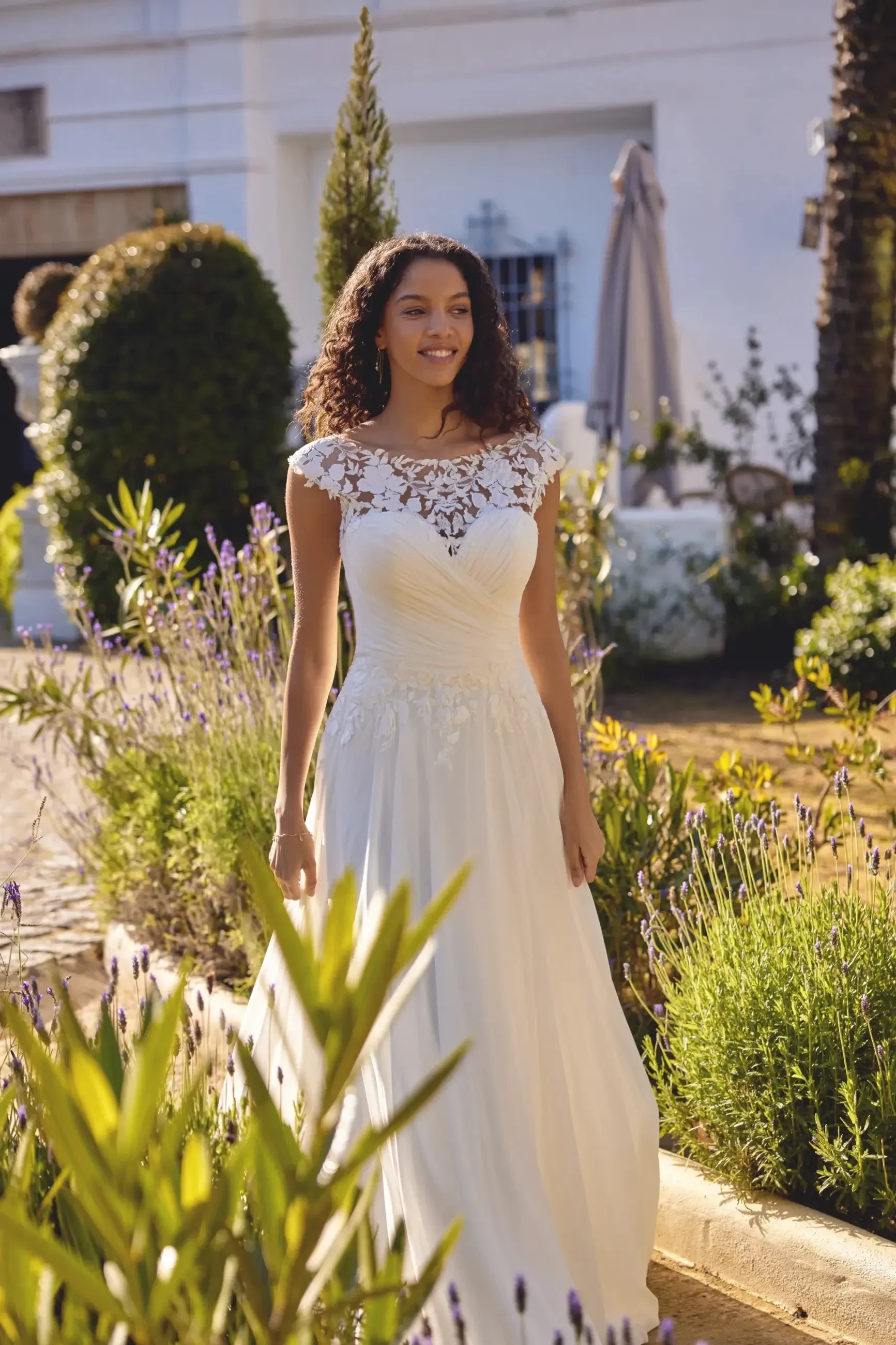 A woman in a white wedding dress with lace detailing, standing outdoors in a garden surrounded by greenery and flowers.