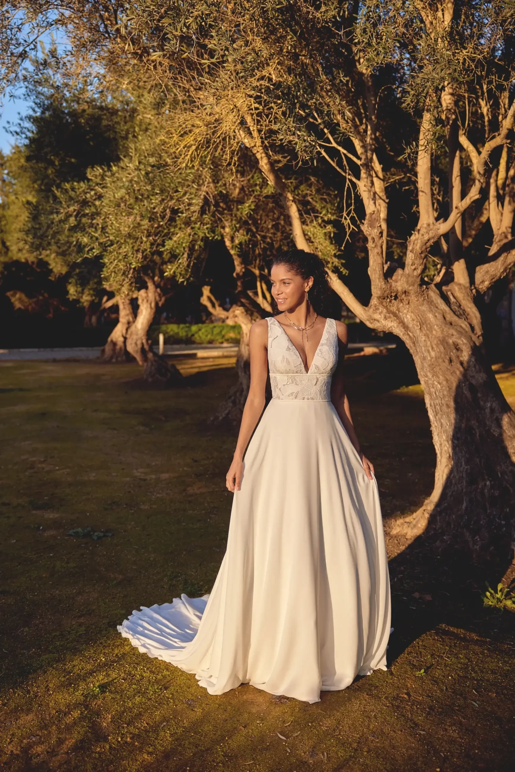 A woman wearing a white bridal gown stands in a natural setting with olive trees, illuminated by warm sunlight.