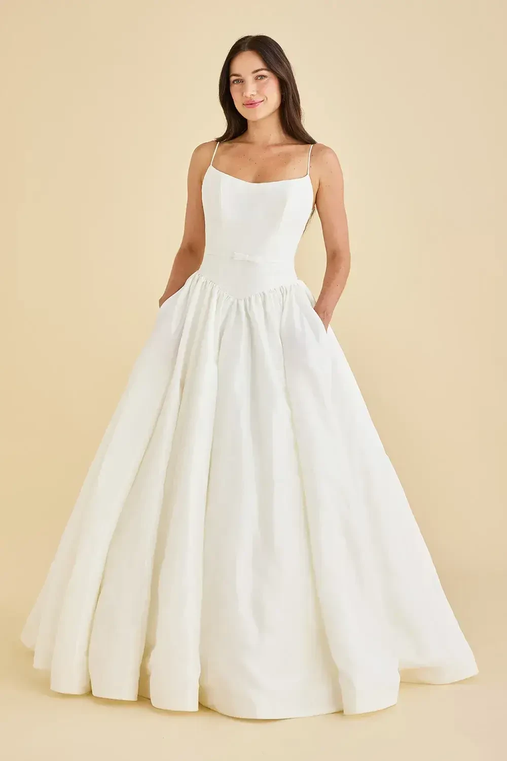 A woman wearing a fitted white gown with a full skirt and pockets, standing against a light beige background.