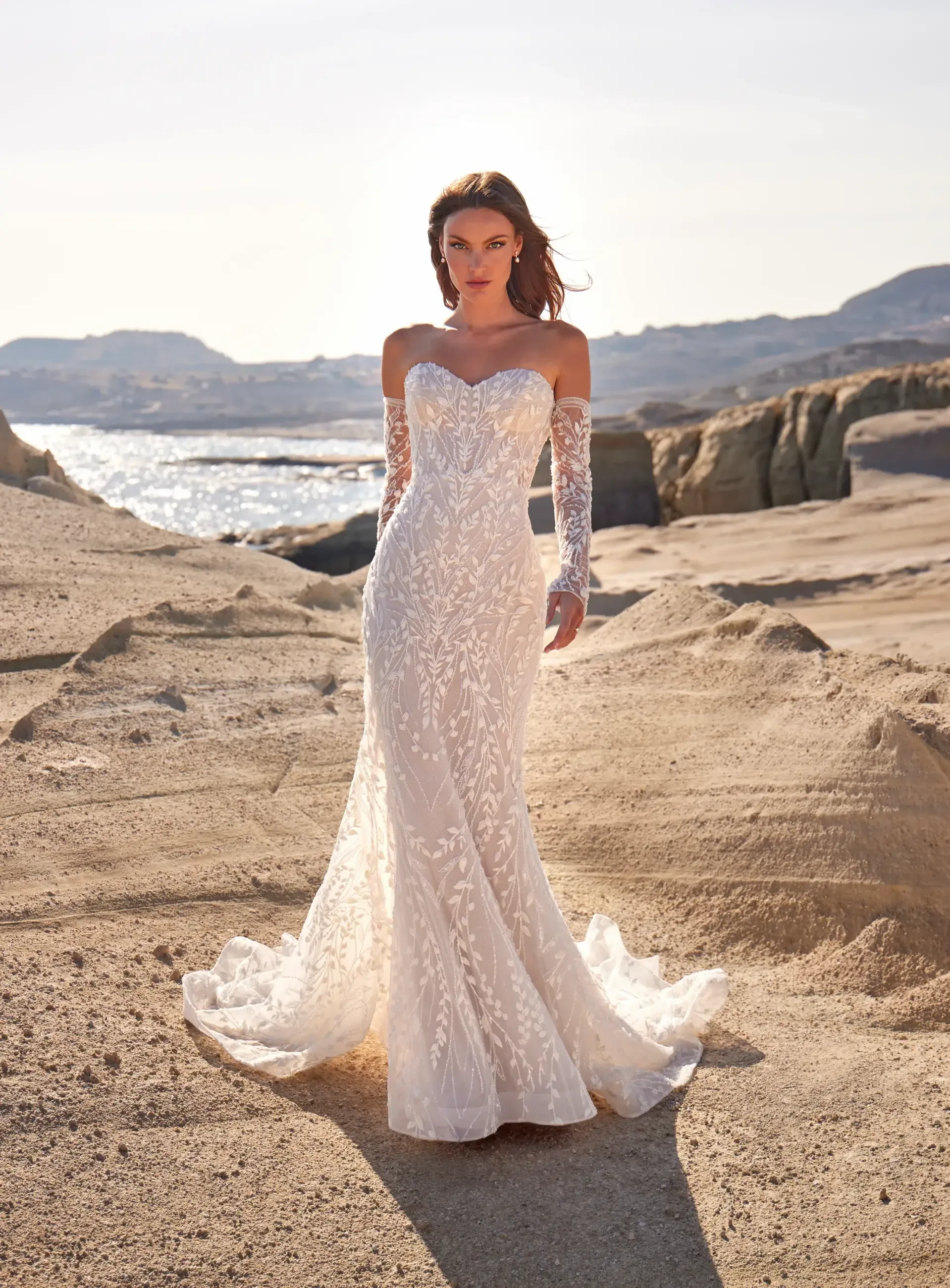 A woman wearing a long, elegant white wedding dress with floral details and long lace sleeves, standing on a beach with rocky cliffs and water in the background.