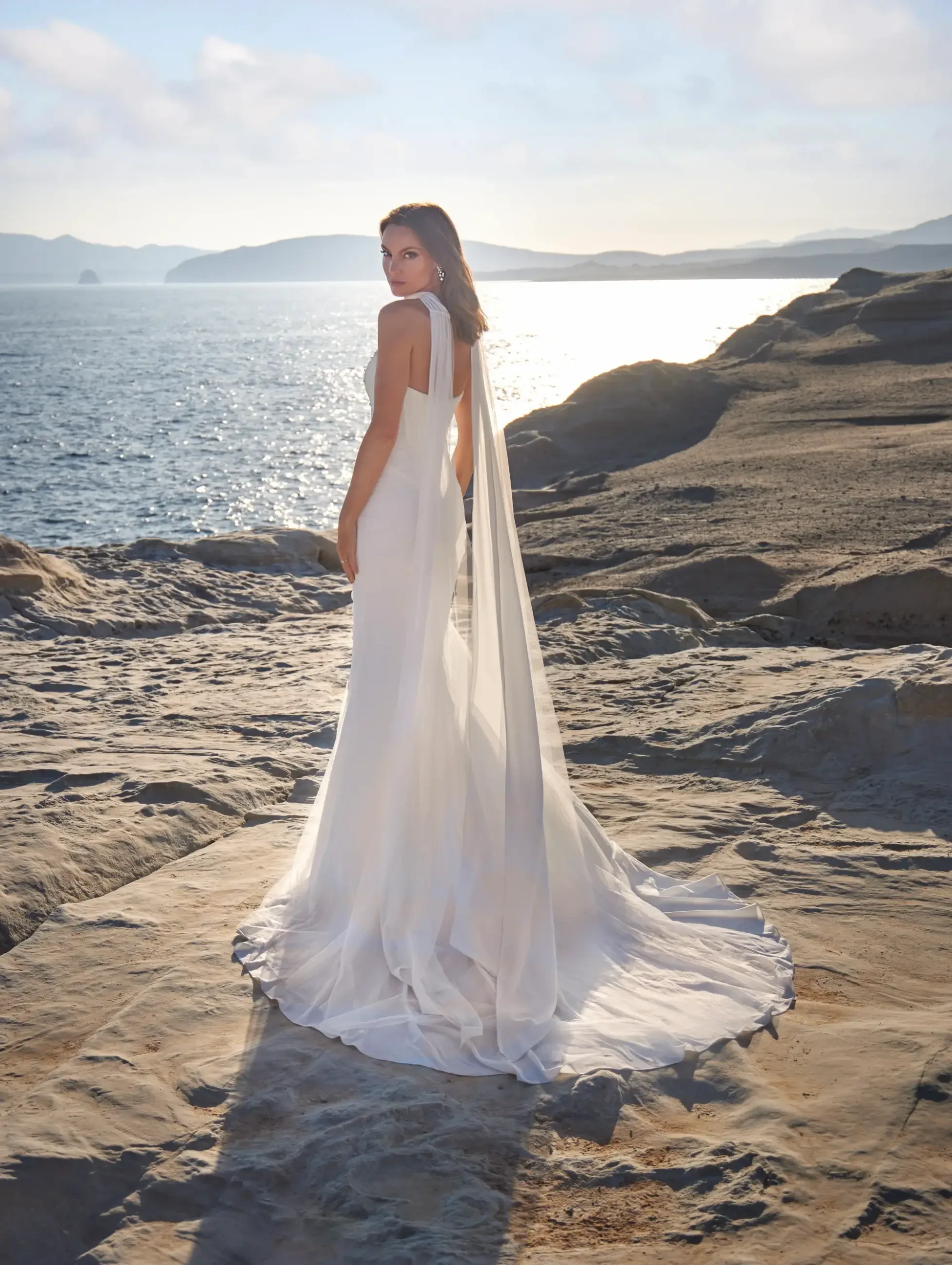 A woman stands on a rocky shoreline wearing a white wedding gown with a long train and veil, looking over her shoulder towards the ocean. The sun is shining softly in the background, creating a serene atmosphere.