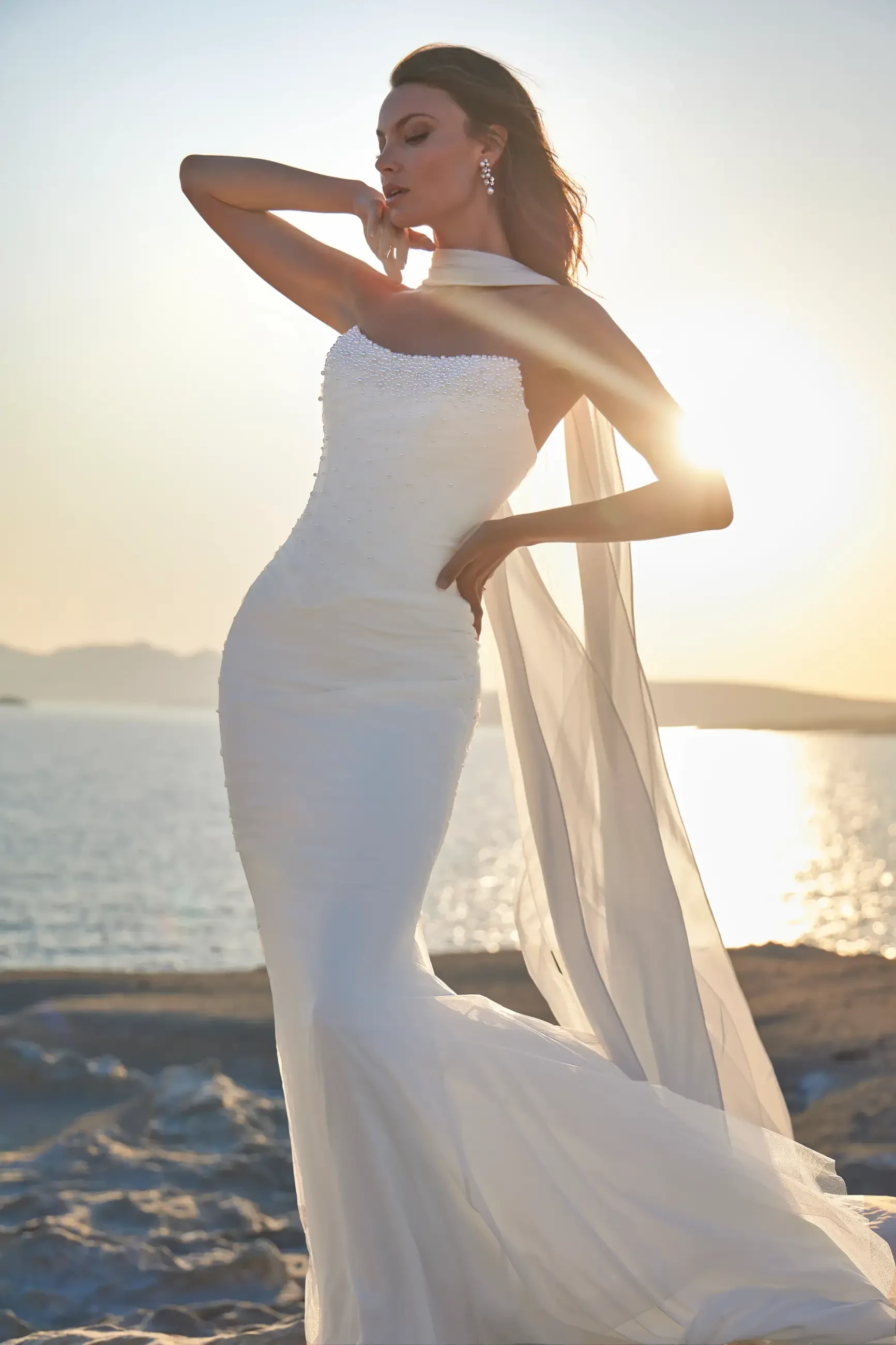 A woman in a white wedding dress poses gracefully against a sunset at the beach. The dress has a fitted silhouette and flowing train, and she is wearing elegant earrings.