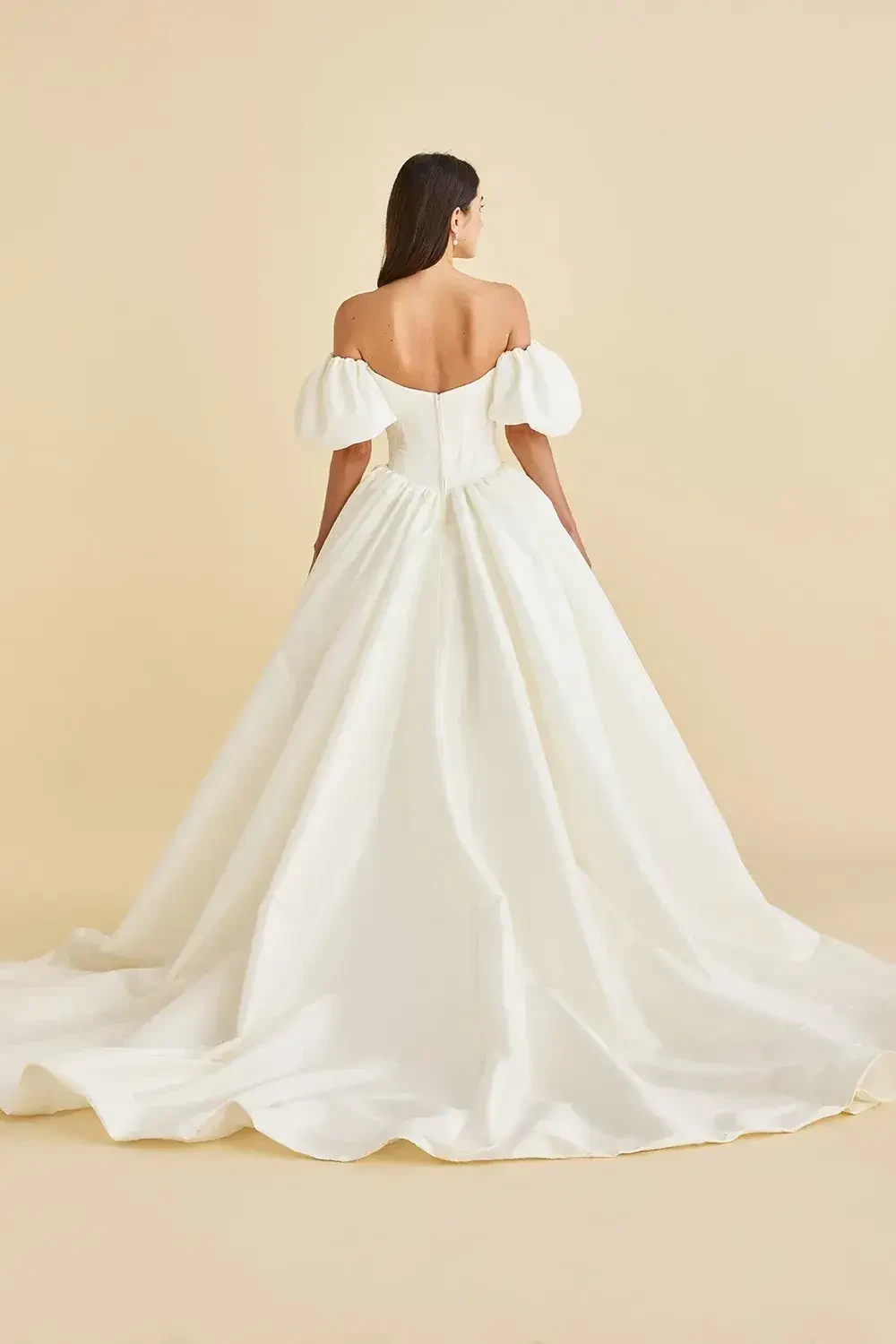 A model wearing a white, off-the-shoulder bridal gown, with a flowing train, shown from the back against a neutral background.