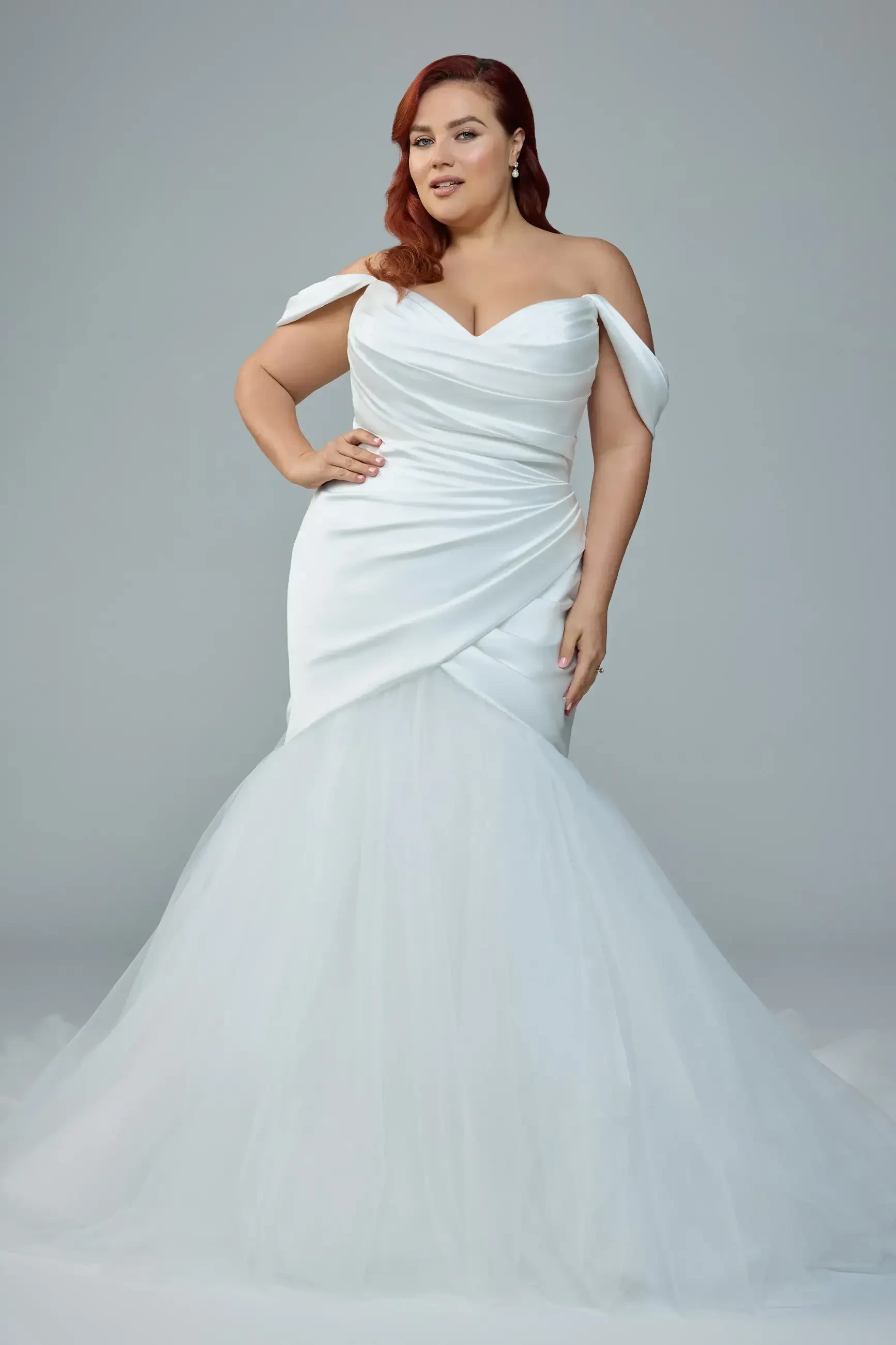 A woman in a white off-shoulder gown with a fitted bodice and a flared skirt. She has wavy red hair and is posing confidently.