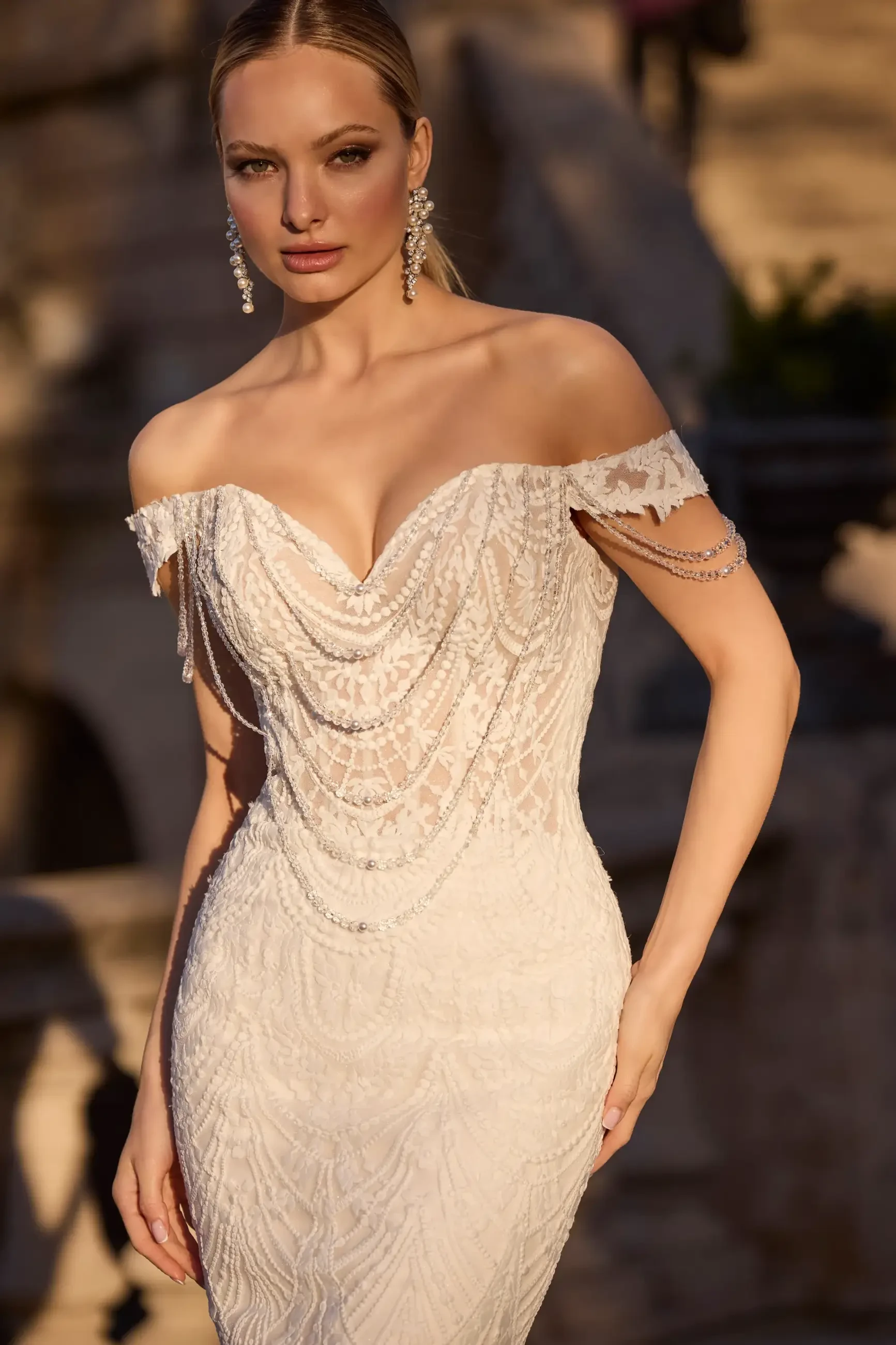 A model in an elegant off-the-shoulder white dress with intricate beadwork, posed outdoors with a soft focus background.