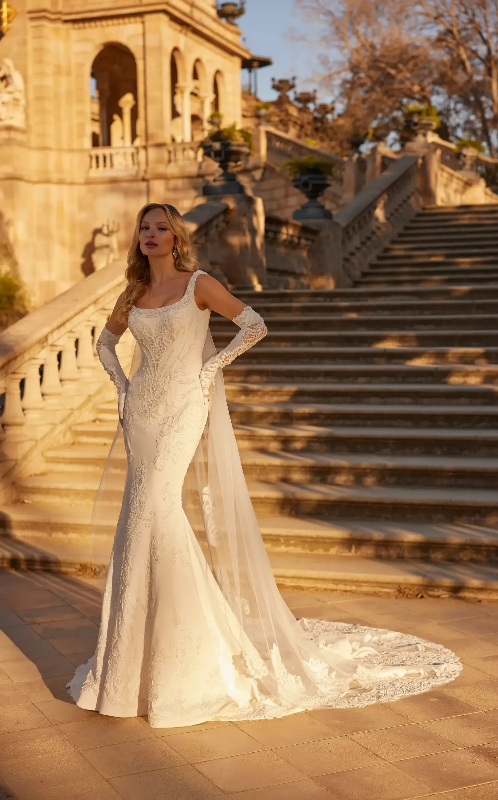 A woman in a white wedding dress with long gloves stands on a staircase in an outdoor setting, illuminated by warm sunlight.