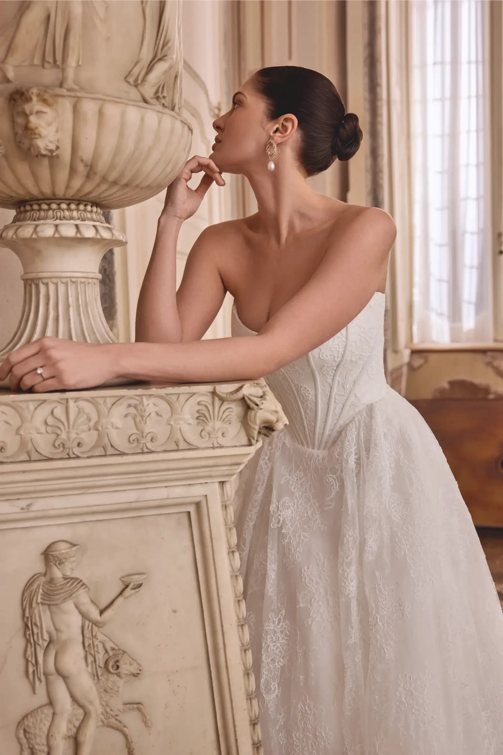 A woman in a strapless white dress poses by an ornate stone structure, with her hand resting on it and looking thoughtfully upward.