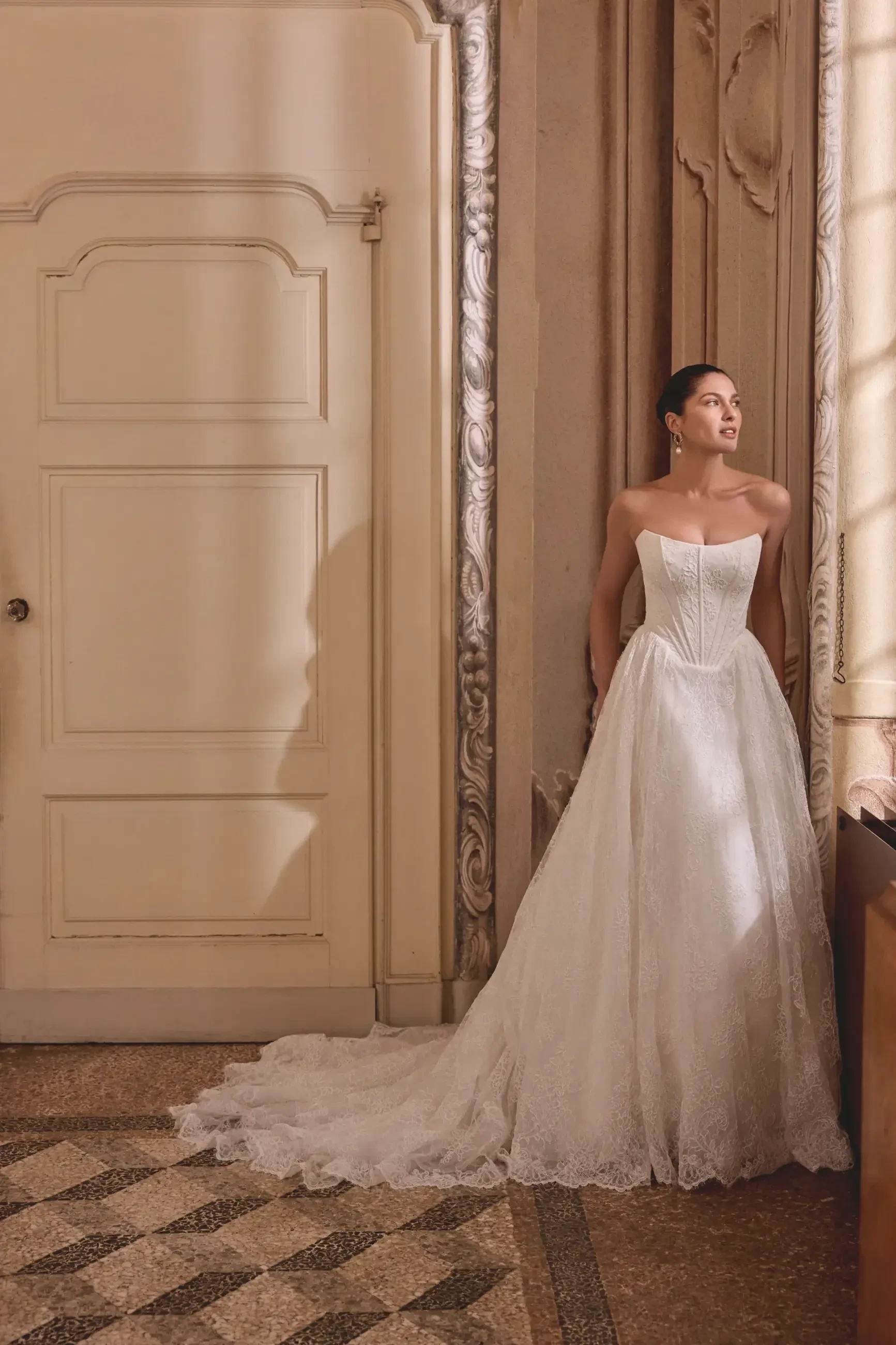 A woman in a white wedding dress stands elegantly next to a wall, with light streaming in from a nearby window, casting shadows on the floor.