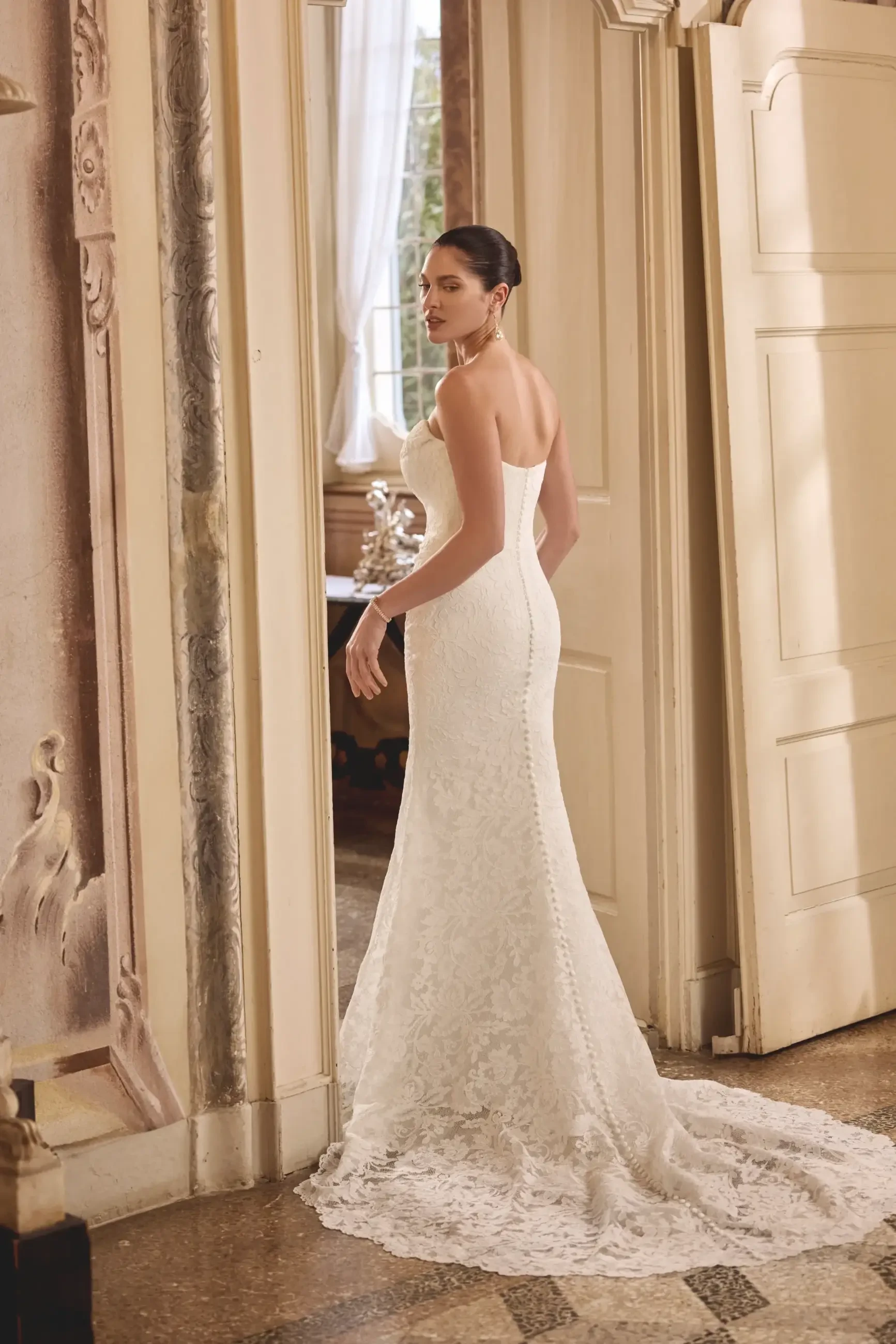 A woman wearing a fitted white wedding dress with lace details, standing near an open doorway in an elegant interior space.