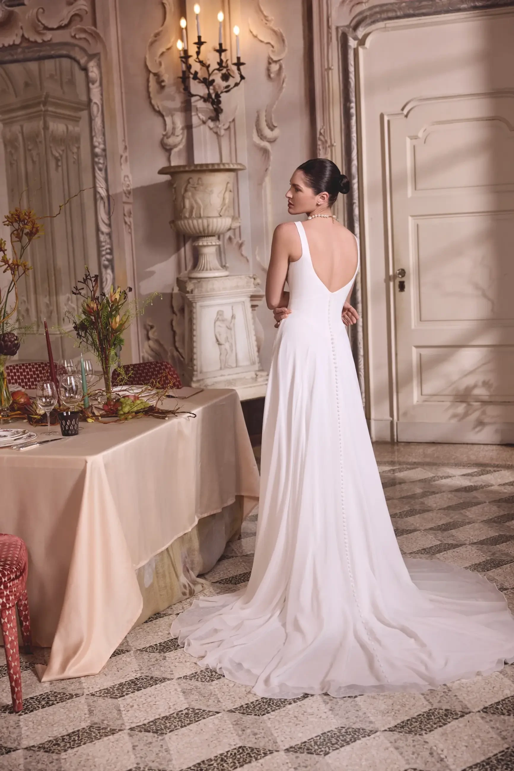 A woman in a white dress stands with her back towards the camera in an elegant room with ornate decor, including a chandelier and a beautiful dining table.