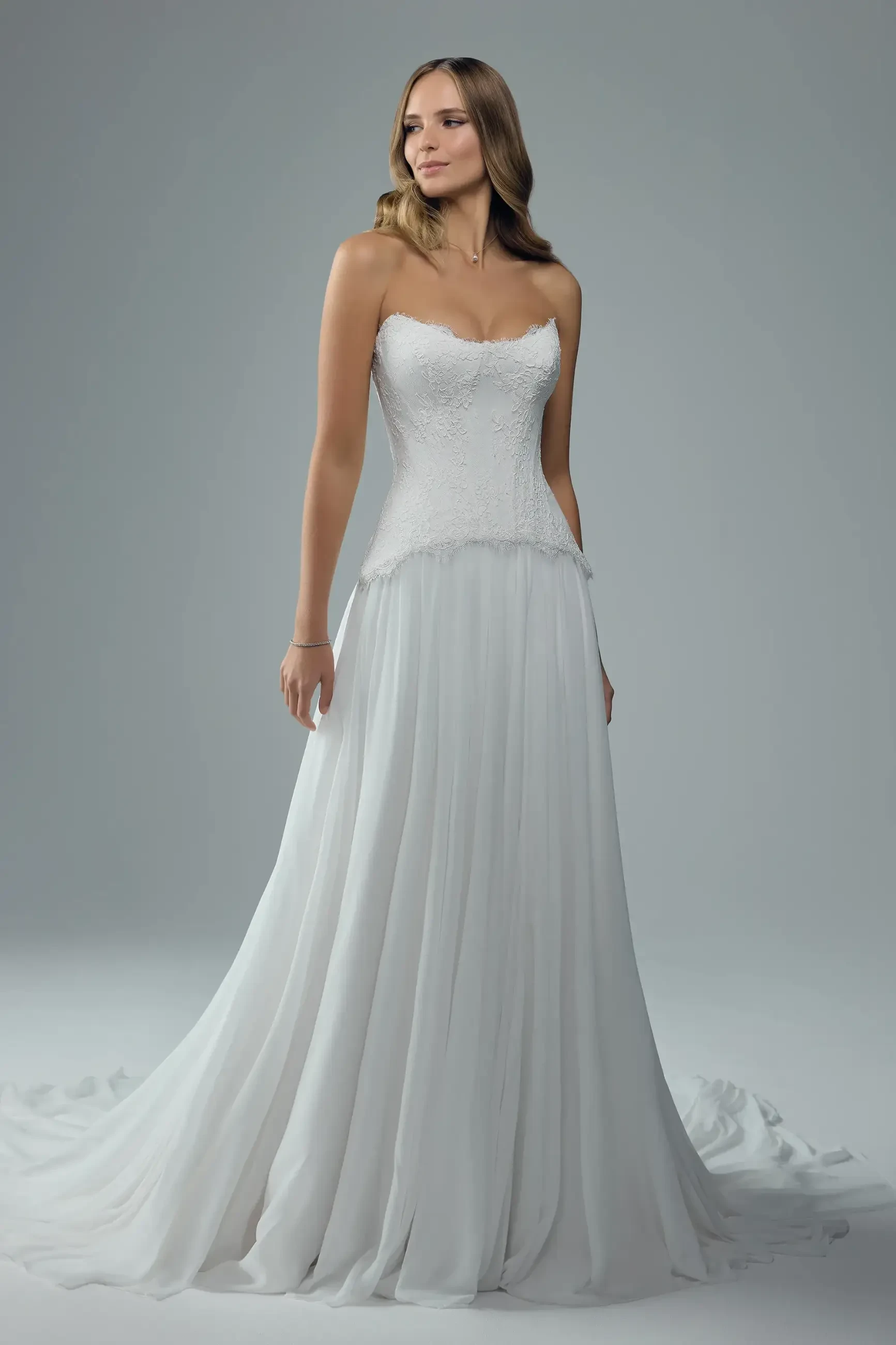 A woman stands gracefully in a strapless white wedding gown with a fitted lace bodice and flowing chiffon skirt, exuding elegance and serenity.